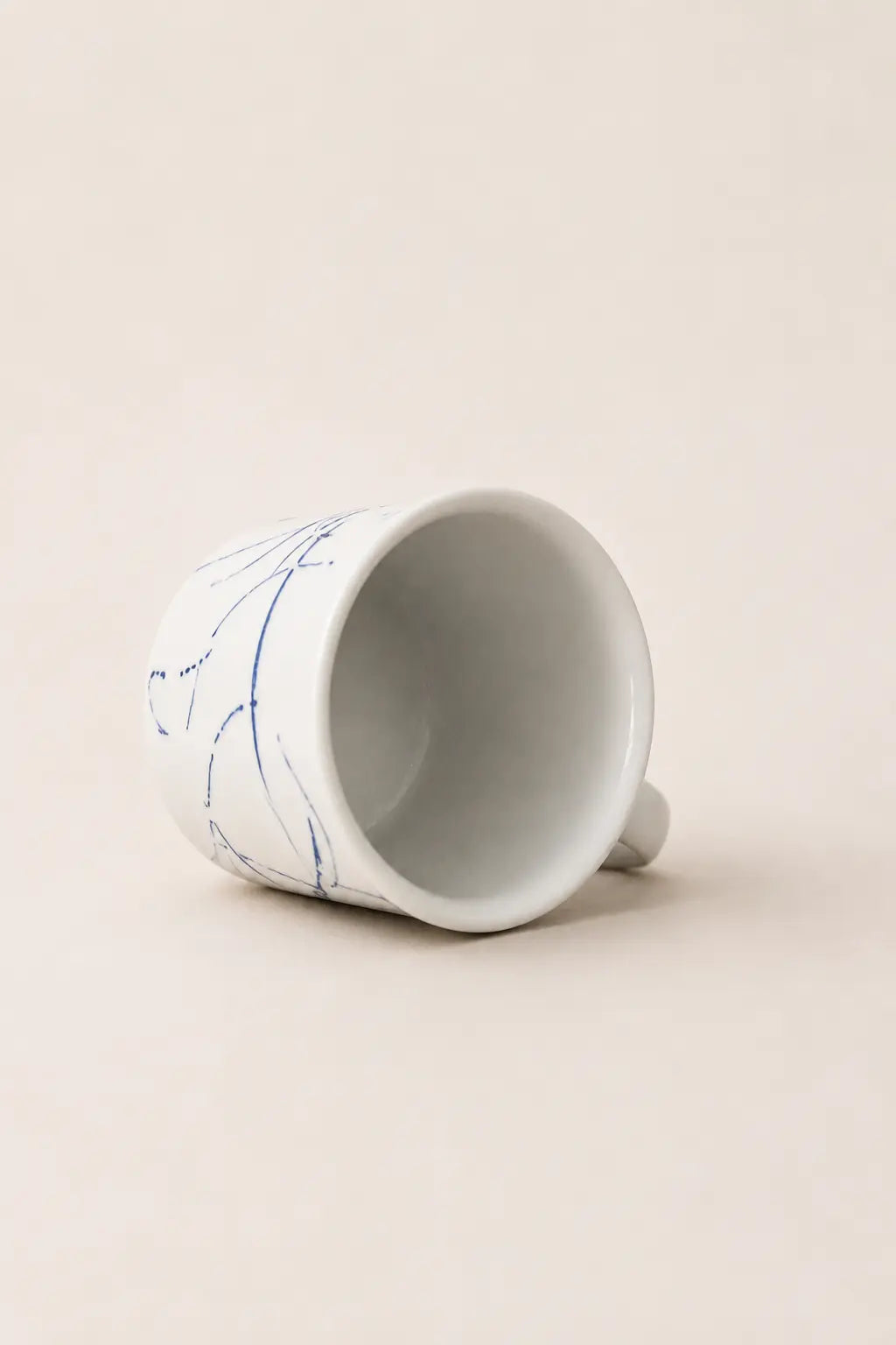 A white ceramic cup with delicate blue hand-painted floral lines, shown lying on its side on a warm beige background.