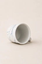 A white ceramic cup with delicate blue hand-painted floral lines, shown lying on its side on a warm beige background.
