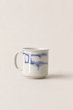 A white ceramic mug with a blue watercolor-style abstract stripe pattern, set against a solid, warm off-white background.