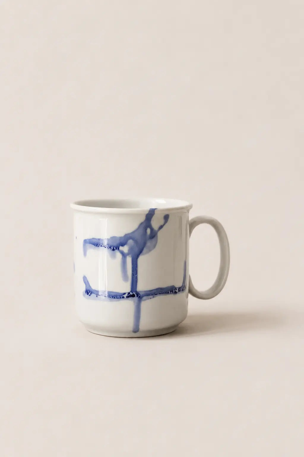 A white ceramic mug with a blue abstract painted design, set against a solid, warm off-white background.