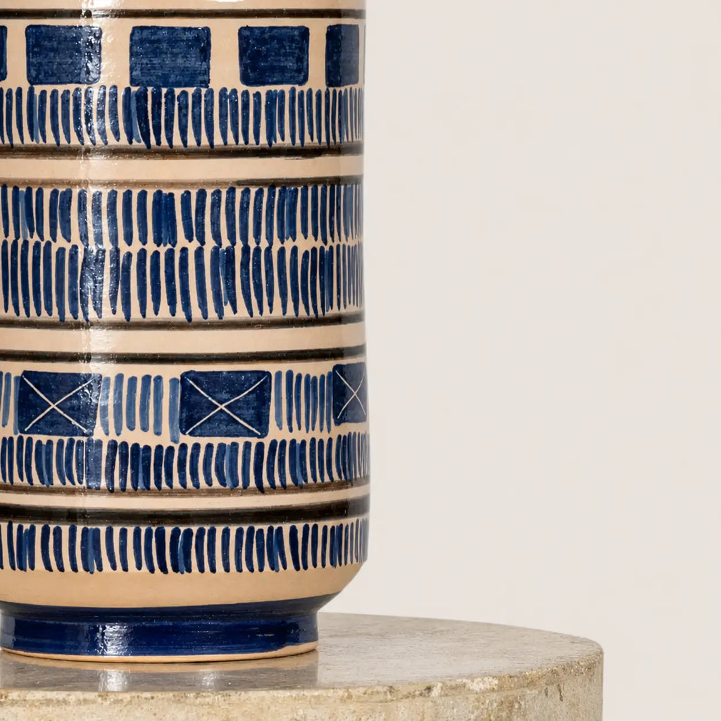 Close-up of a ceramic vase with a cream-colored glaze and dark blue geometric patterns, displayed on a round stone pedestal against a light background.