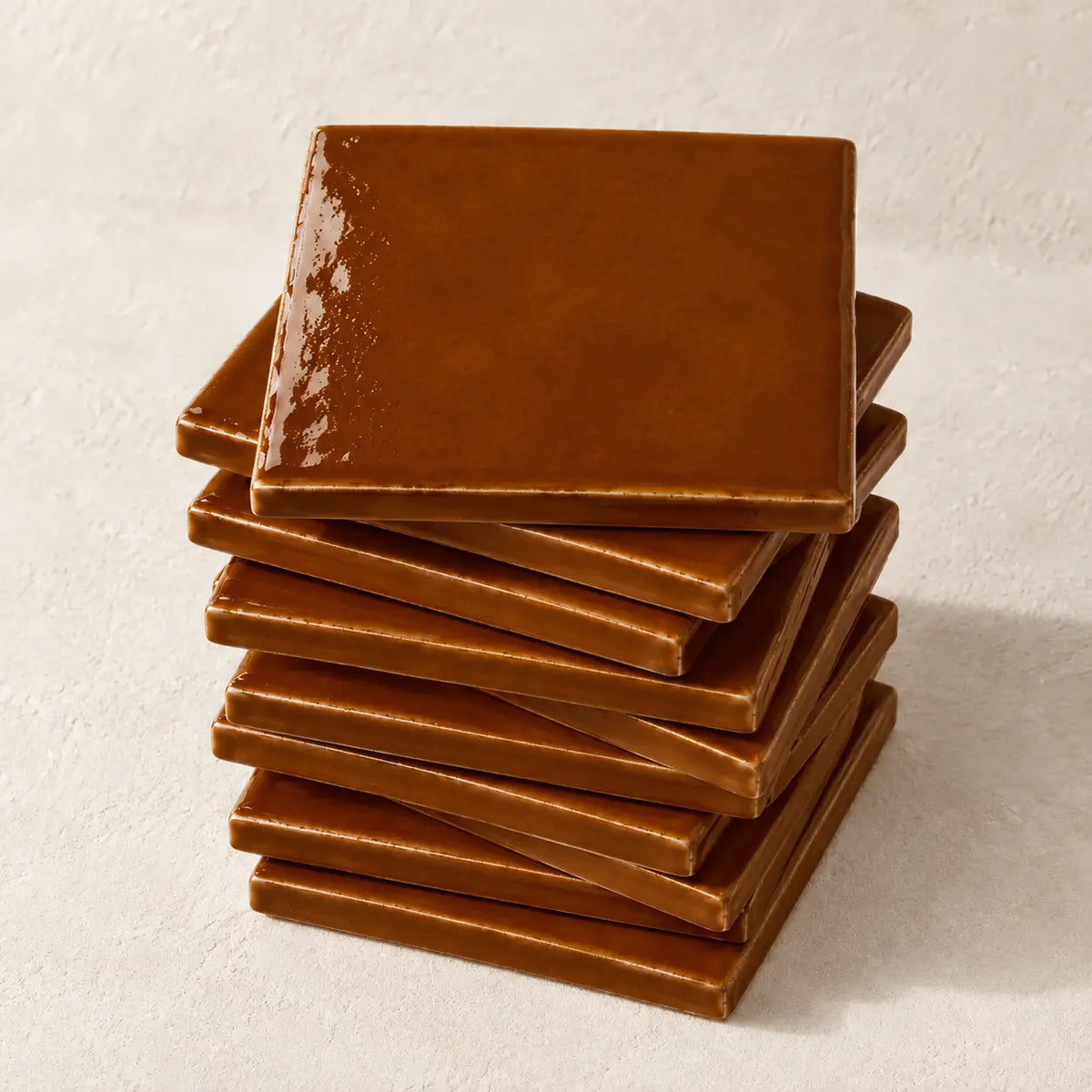 A stack of glossy square ceramic tiles in a deep cognac brown colour, arranged on a textured off-white surface.