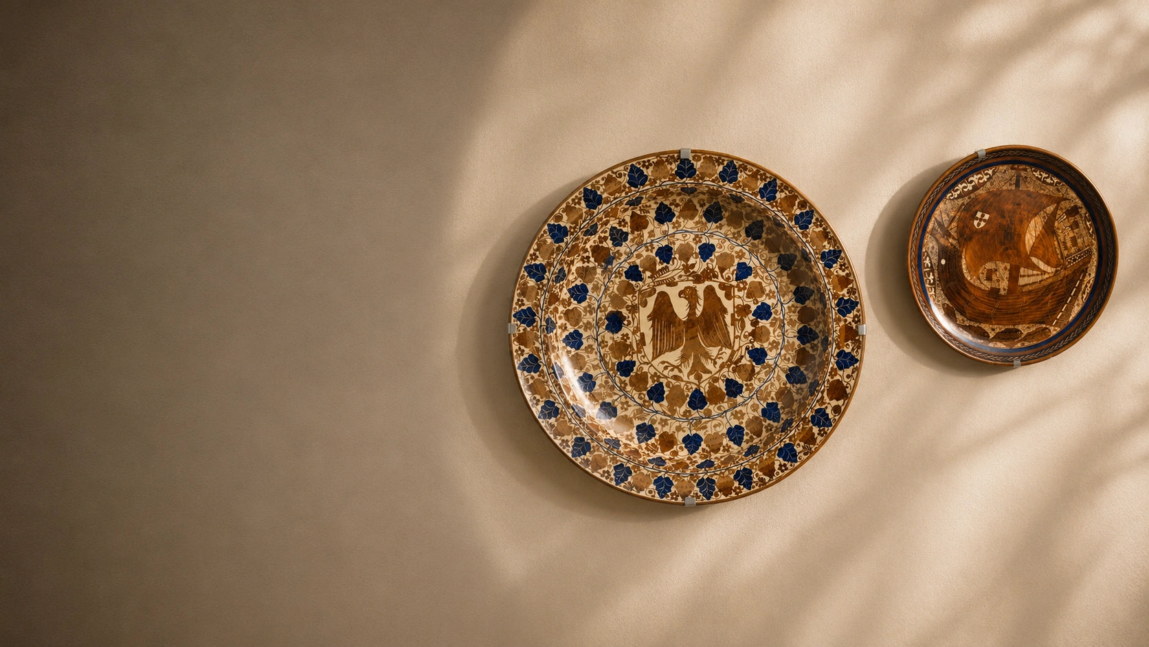 Two decorative ceramic plates with intricate blue and gold patterns, displayed on a warm beige wall with soft, dappled sunlight casting shadows.