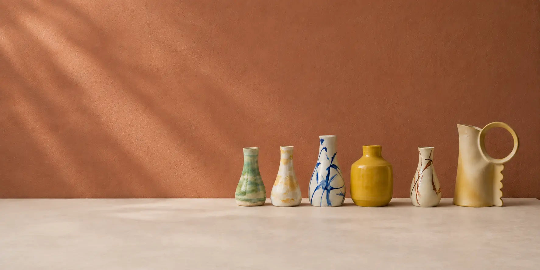 Six small ceramic vases and a matching pitcher arranged in a row on a light surface against a warm terracotta background. The vessels have varied glazes and patterns in muted tones including green, cream, blue, yellow, and beige.