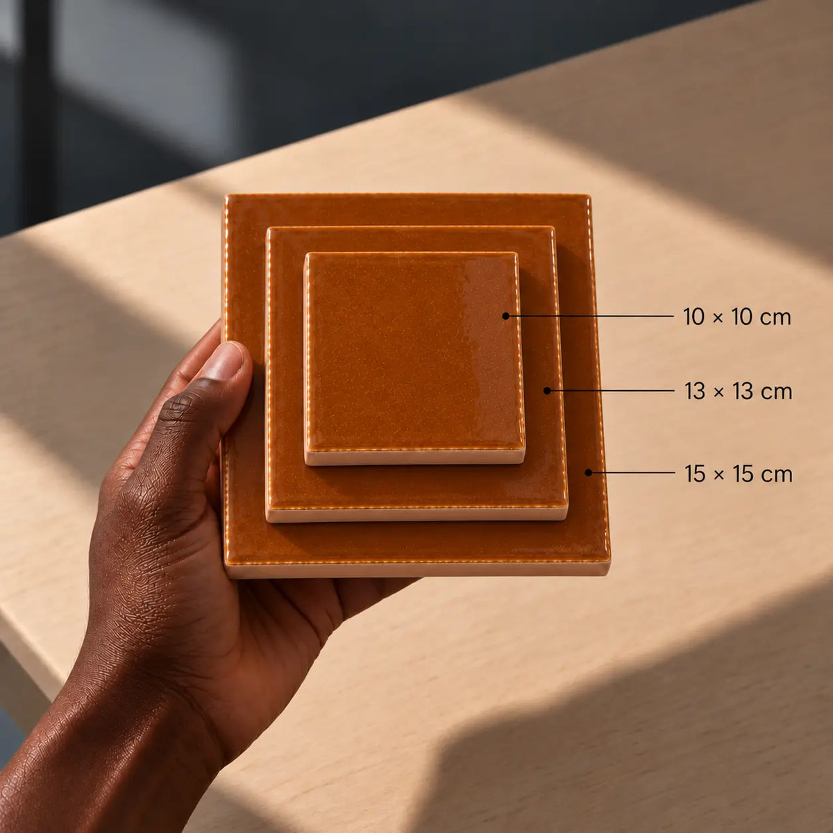 A hand holds a stack of three glossy square ceramic tiles in warm tan-brown glaze, with size labels pointing to 10 x 10 cm, 13 x 13 cm, and 15 x 15 cm over a light wood tabletop.