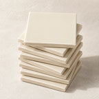 A stack of square ceramic coasters in a warm off-white cream color, arranged neatly on a textured light beige surface.