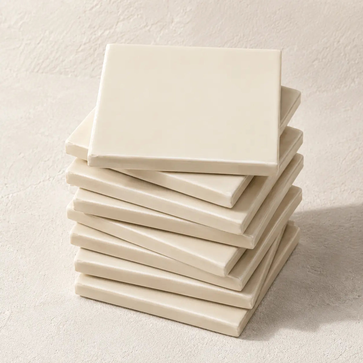 A stack of square ceramic coasters in a warm off-white cream color, arranged neatly on a textured light beige surface.