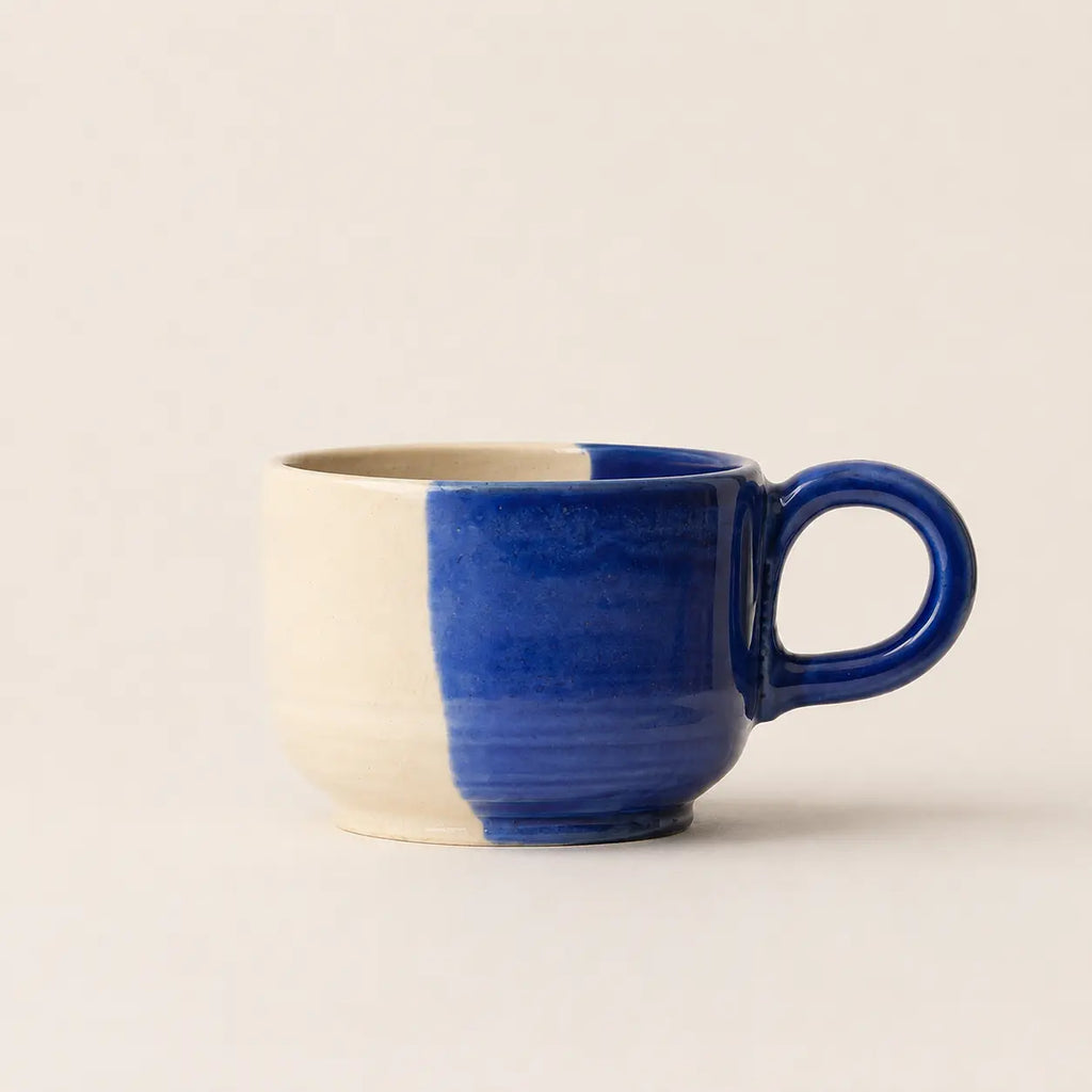A ceramic coffee mug with a vertical split design, featuring a matte cream-colored left side and a glossy deep cobalt blue right side and handle, set against a plain cream background.