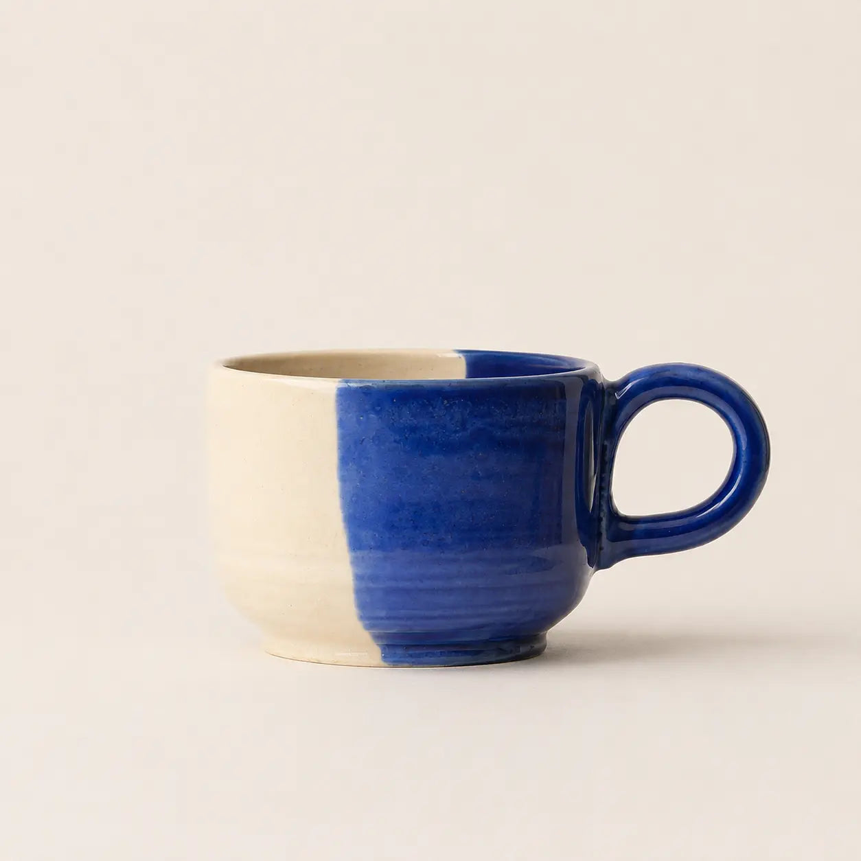 A ceramic coffee mug with a vertical split design, featuring a matte cream-colored left side and a glossy deep cobalt blue right side and handle, set against a plain cream background.