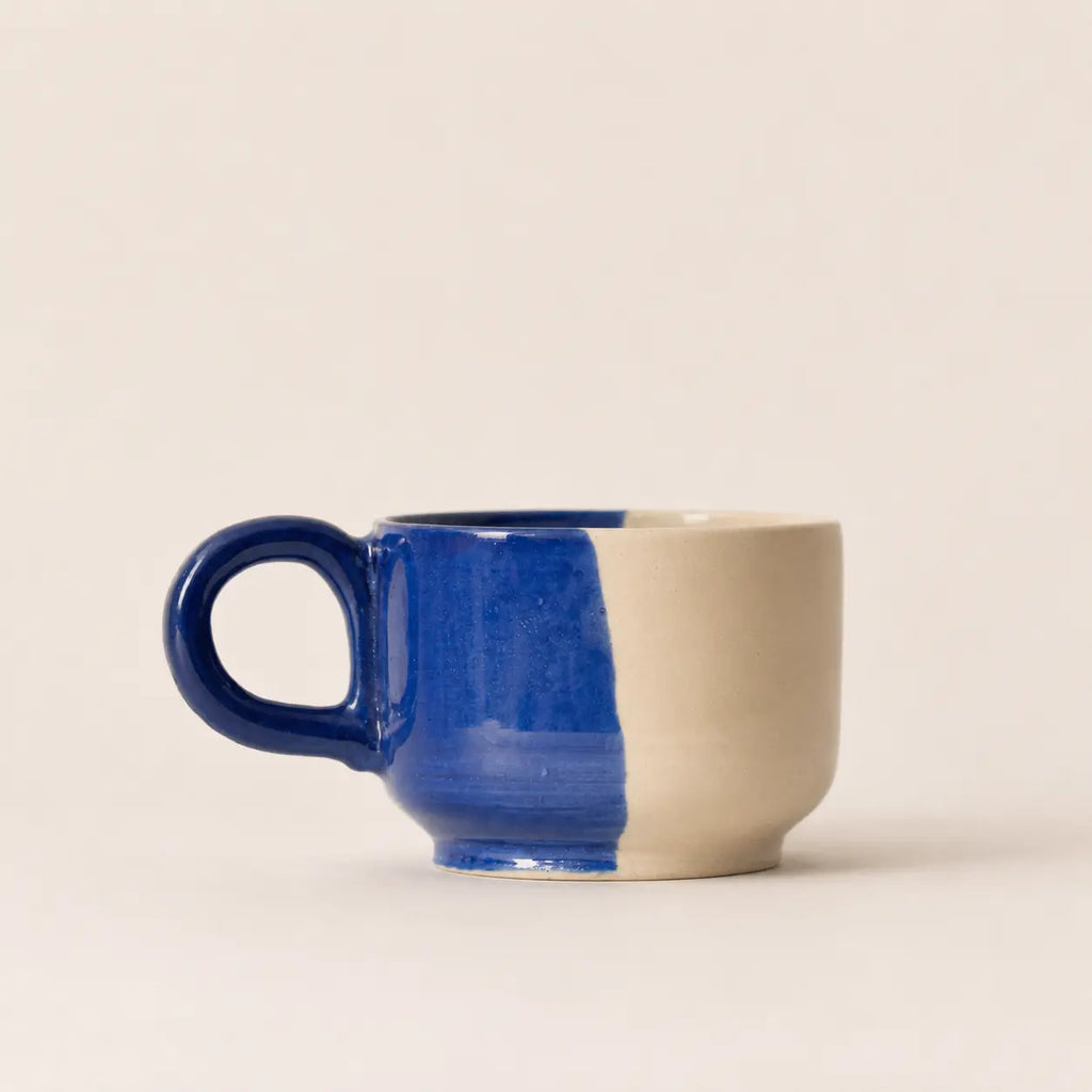 A ceramic coffee cup with a split blue and beige glaze, featuring a dark blue handle and rim, set against a plain beige background.