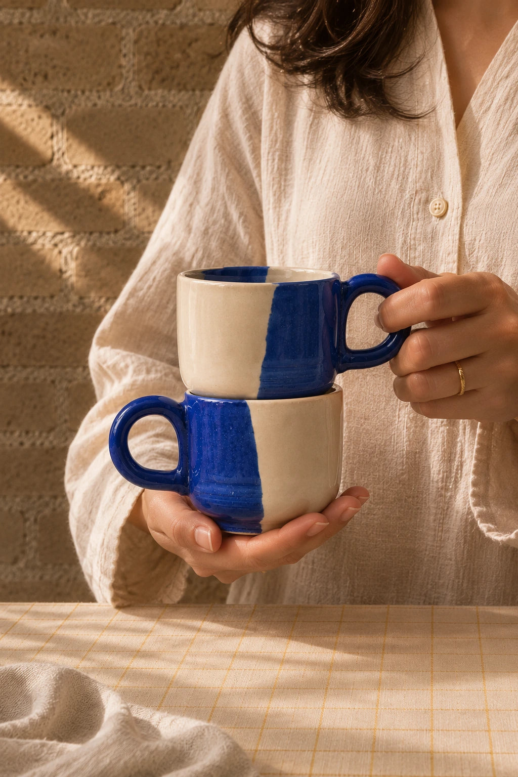 A person in a cream linen shirt holds two stacked ceramic mugs with a bold blue and cream color-blocked design, set against a warm, textured brick wall with soft window light.