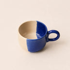 A ceramic coffee cup with a split-color glaze, featuring a deep cobalt blue exterior and handle, and a warm cream-beige interior, set against a solid light beige background.