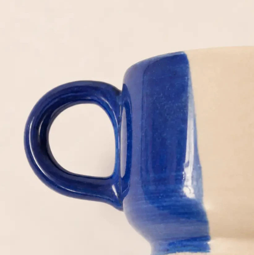 Close-up of a ceramic coffee mug showing a glossy cobalt blue handle and upper body with a warm cream-colored lower section, set against a plain light beige background.