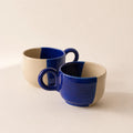 Two ceramic coffee mugs with a two-tone design of deep cobalt blue and warm cream, arranged on a light beige background.