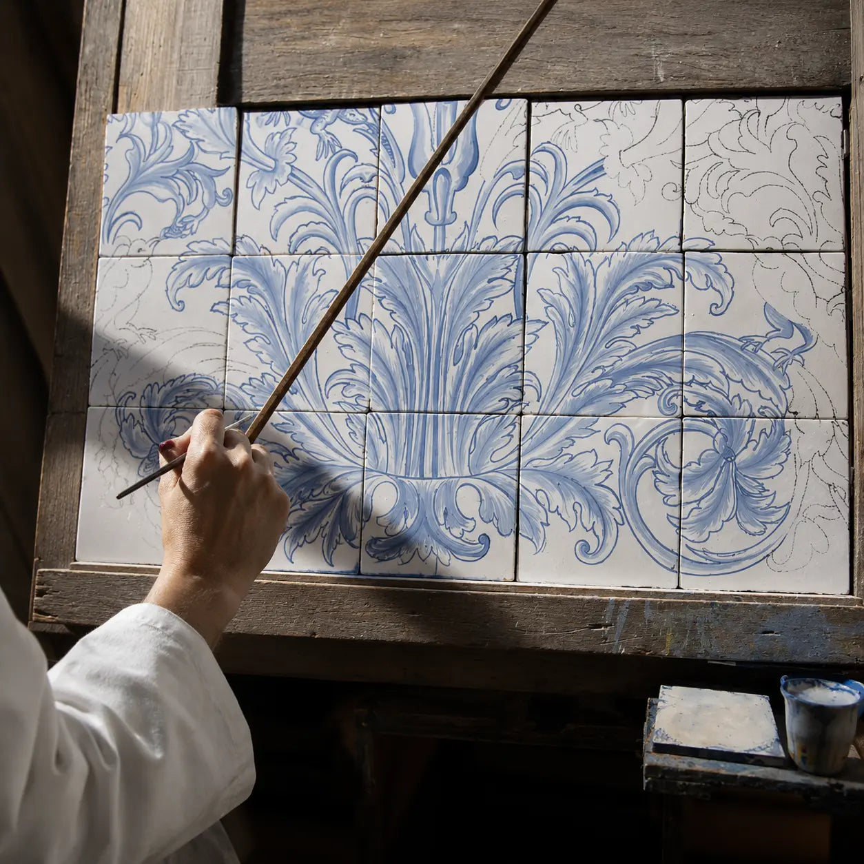 A hand holding a paintbrush applies blue floral detailing to a set of white ceramic tiles mounted on a rustic wooden panel.