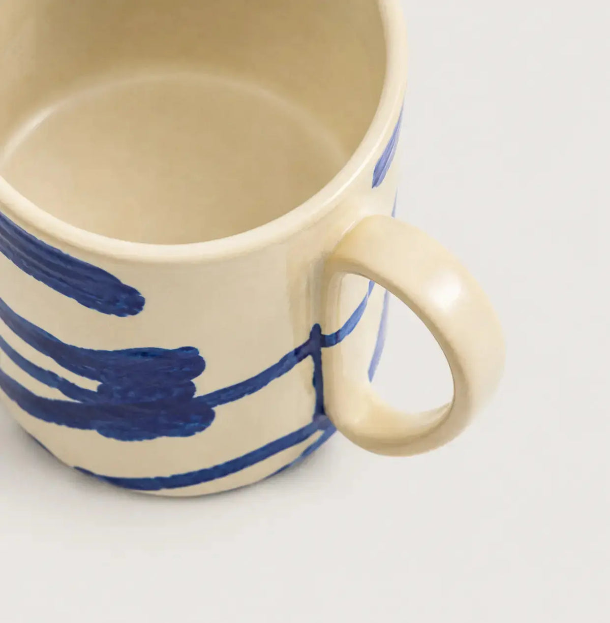 A close-up of a porcelain mug with a cream-colored glaze and bold blue painted brushstroke patterns, shown on a plain white background.