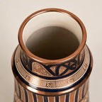 Close-up of a decorative ceramic vase with a copper-toned rim, dark blue and gold patterned band, and vertical fluted stripes on a light neutral background.