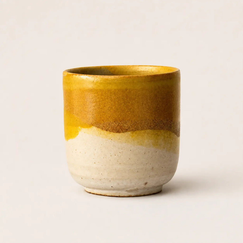 A handmade ceramic espresso cup with a glossy mustard yellow glaze on the upper half and a speckled, unglazed off-white base on the lower half, set against a plain light beige background.