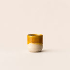 A handmade ceramic espresso cup with a glossy mustard yellow glaze on the upper half and a speckled, unglazed off-white base on the lower half, set against a plain light beige background.