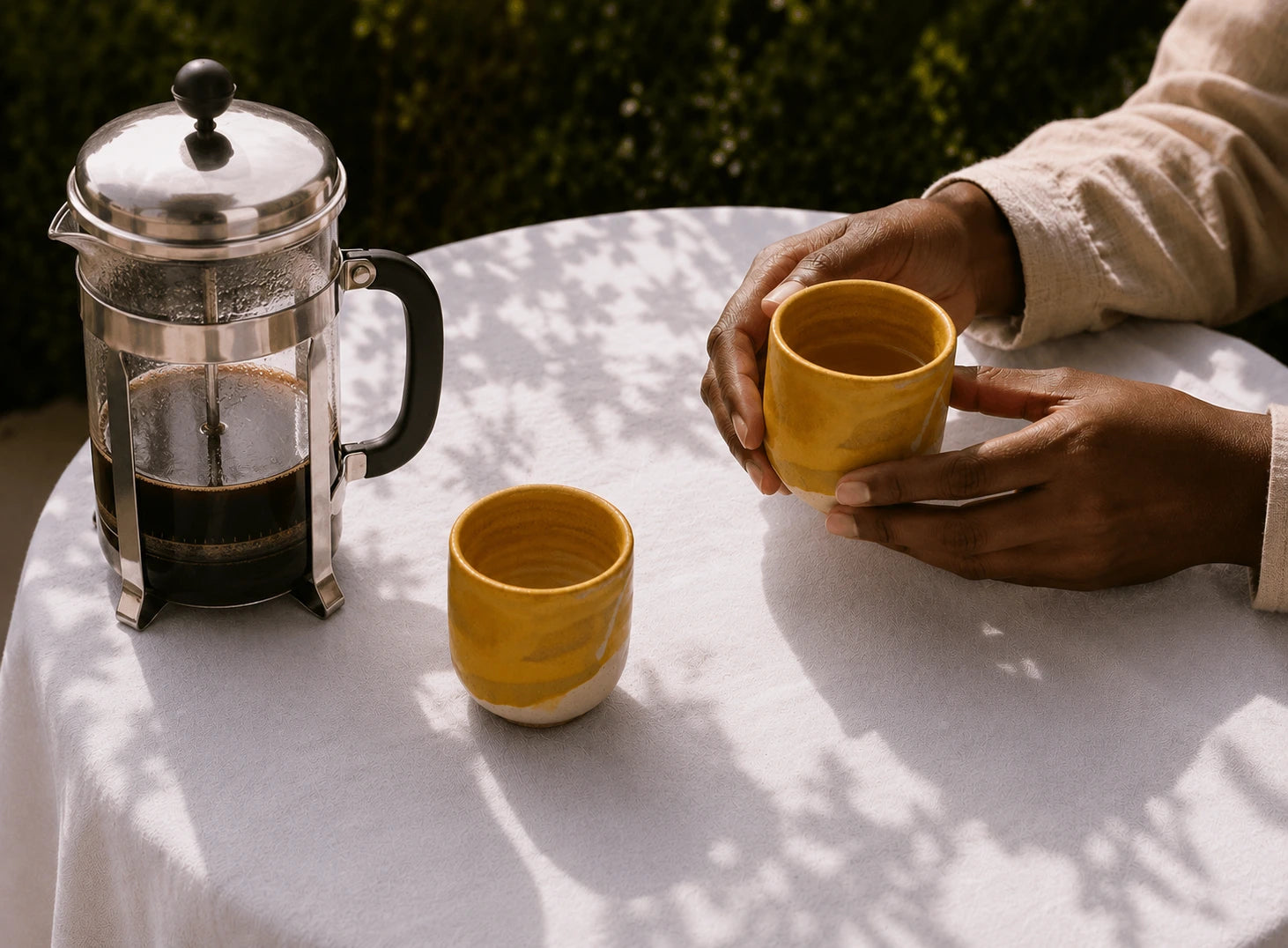 Two yellow ceramic cups on a white tablecloth next to a glass French press, with a person holding one cup in an outdoor setting with dappled sunlight.