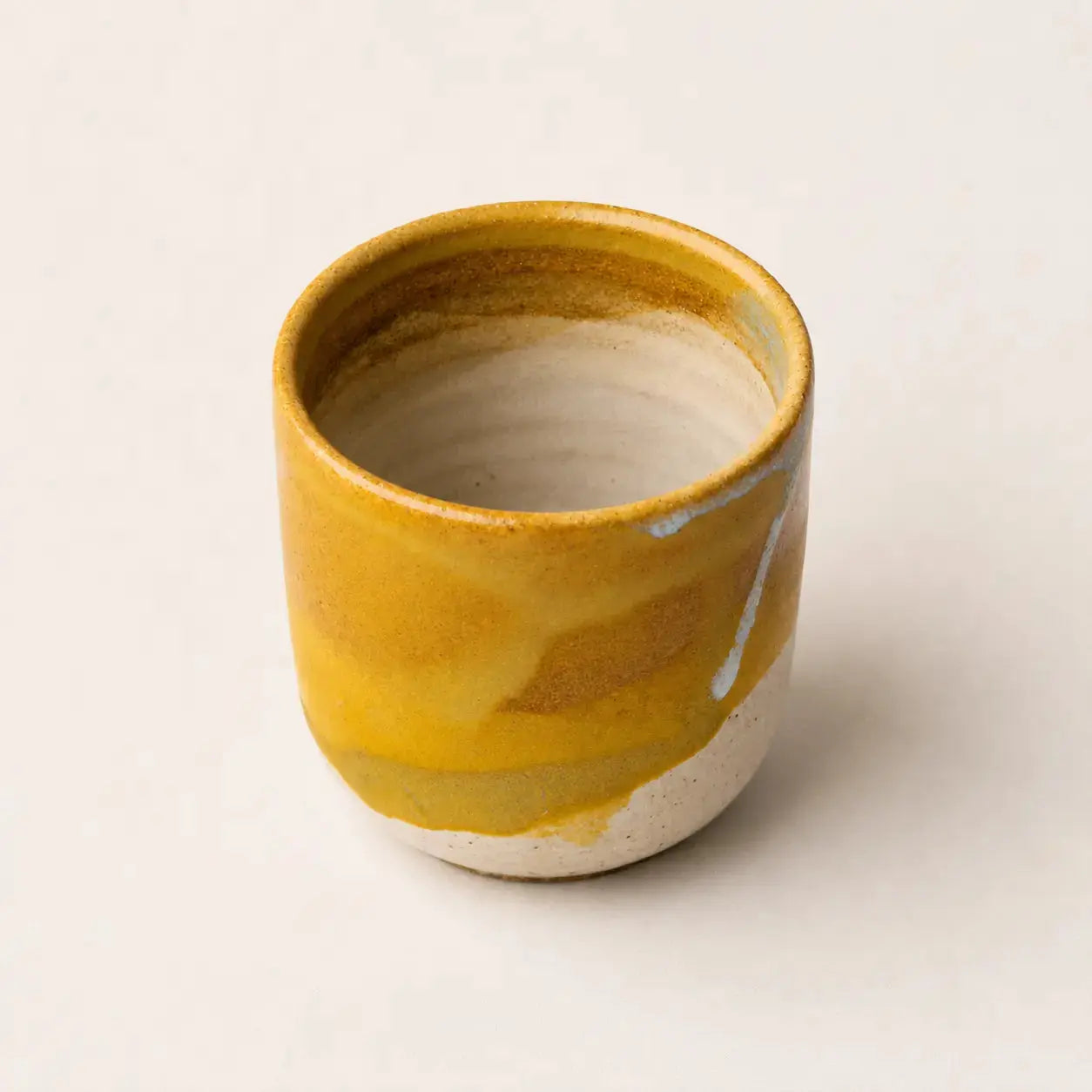 A small handmade ceramic espresso cup with a yellow and ochre glazed exterior and a natural off-white unglazed base, photographed on a light neutral background.
