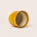 A small handmade ceramic espresso cup with a wide open top, featuring a textured mustard yellow glazed exterior and a matte beige interior, set against a plain off-white background.