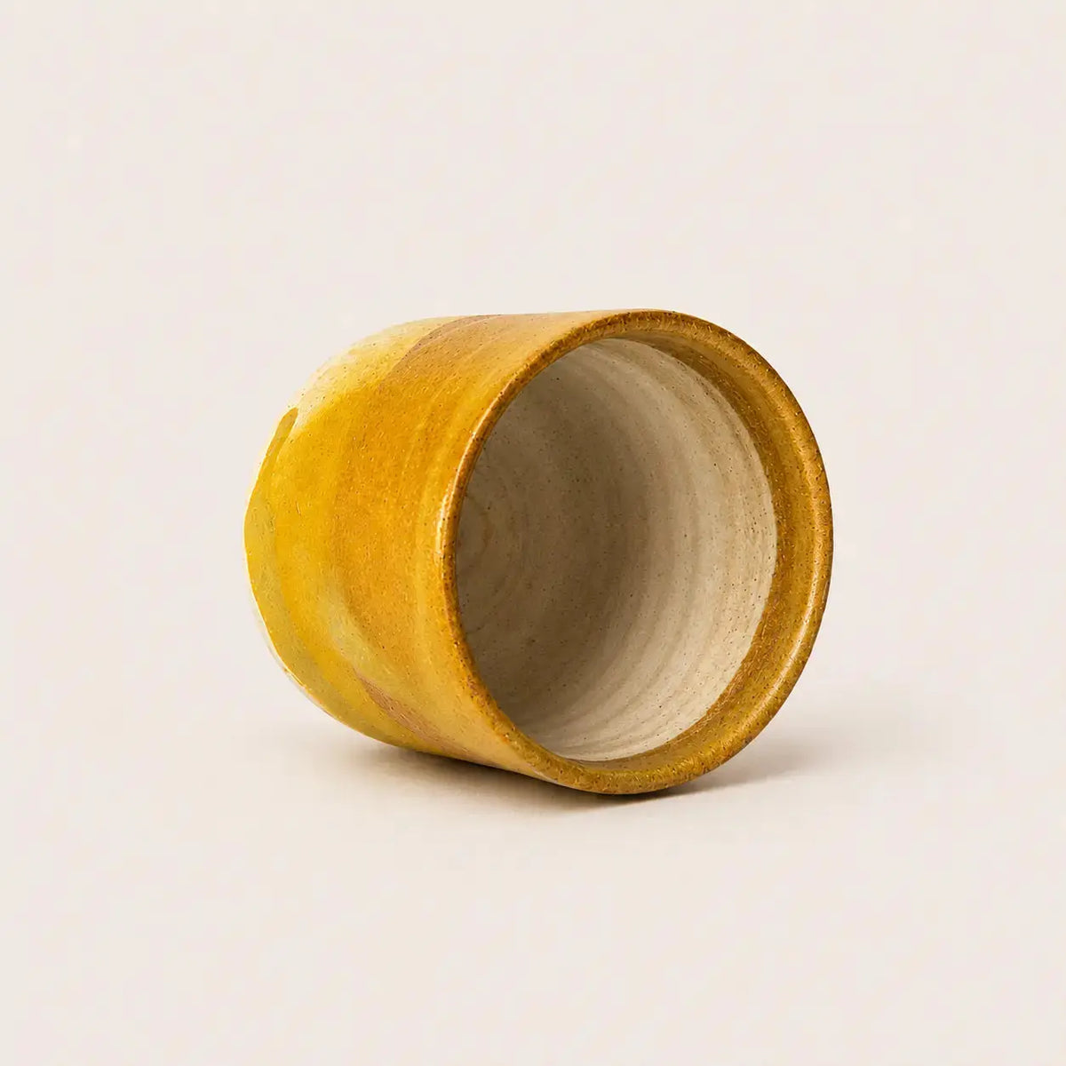 A small handmade ceramic espresso cup with a wide open top, featuring a textured mustard yellow glazed exterior and a matte beige interior, set against a plain off-white background.