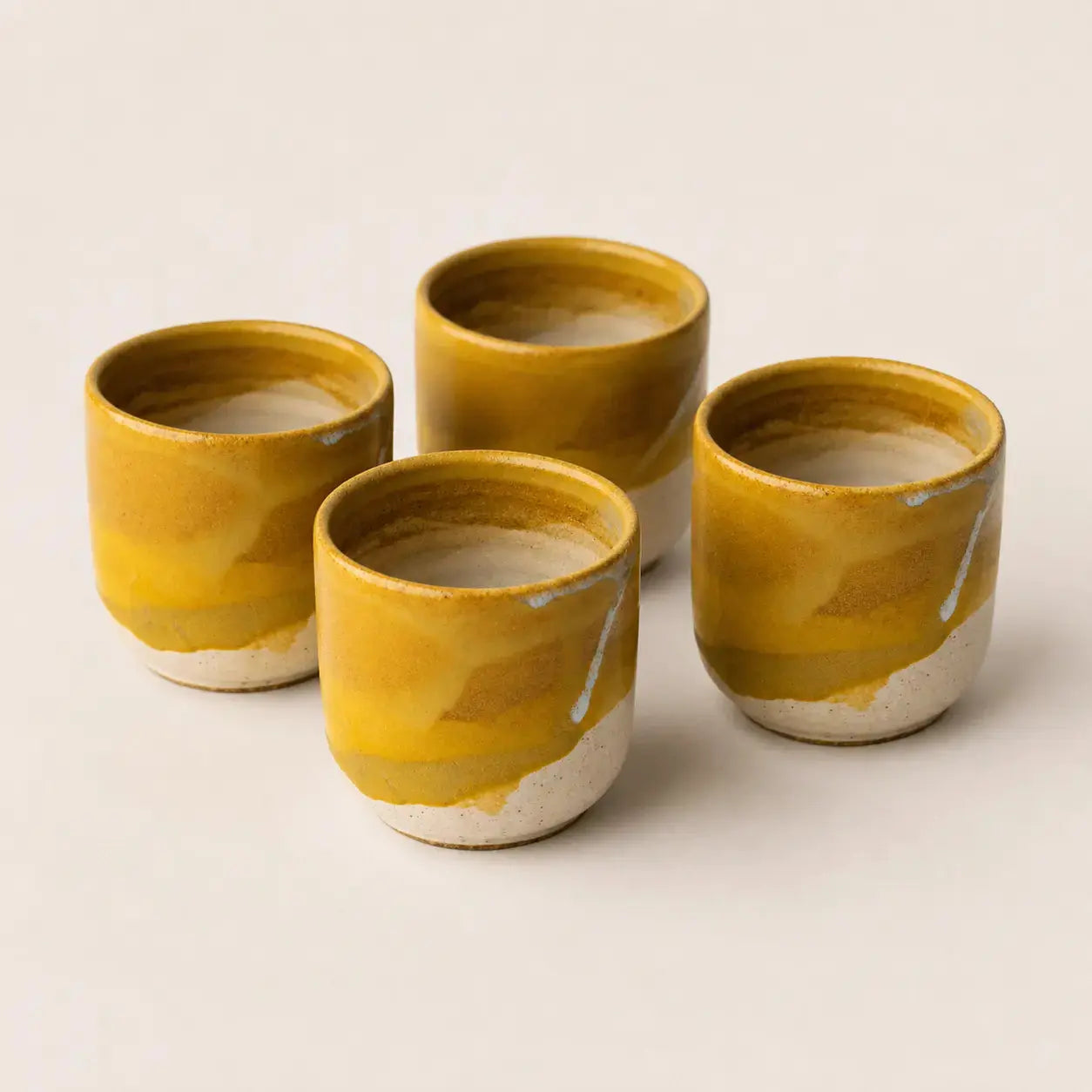 Four small handmade ceramic espresso cup with a marbled mustard yellow and white glaze, arranged on a light beige background.