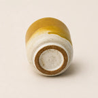 A small handmade ceramic espresso cup shown from the bottom, featuring a glossy mustard yellow glaze on the upper half and a speckled off-white clay body on the lower half, with a natural brown clay base ring.