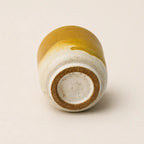 A small handmade ceramic espresso cup shown from the bottom, featuring a glossy mustard yellow glaze on the upper half and a speckled off-white clay body on the lower half, with a natural brown clay base ring.