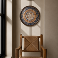A styled interior scene with a wooden armchair in the foreground and two decorative wall plates hanging above it against a beige wall, with sunlight streaming from a window on the left.
