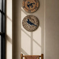 Two decorative circular wall clocks with bird motifs hang on a beige wall above a wooden armchair in a sunlit room.