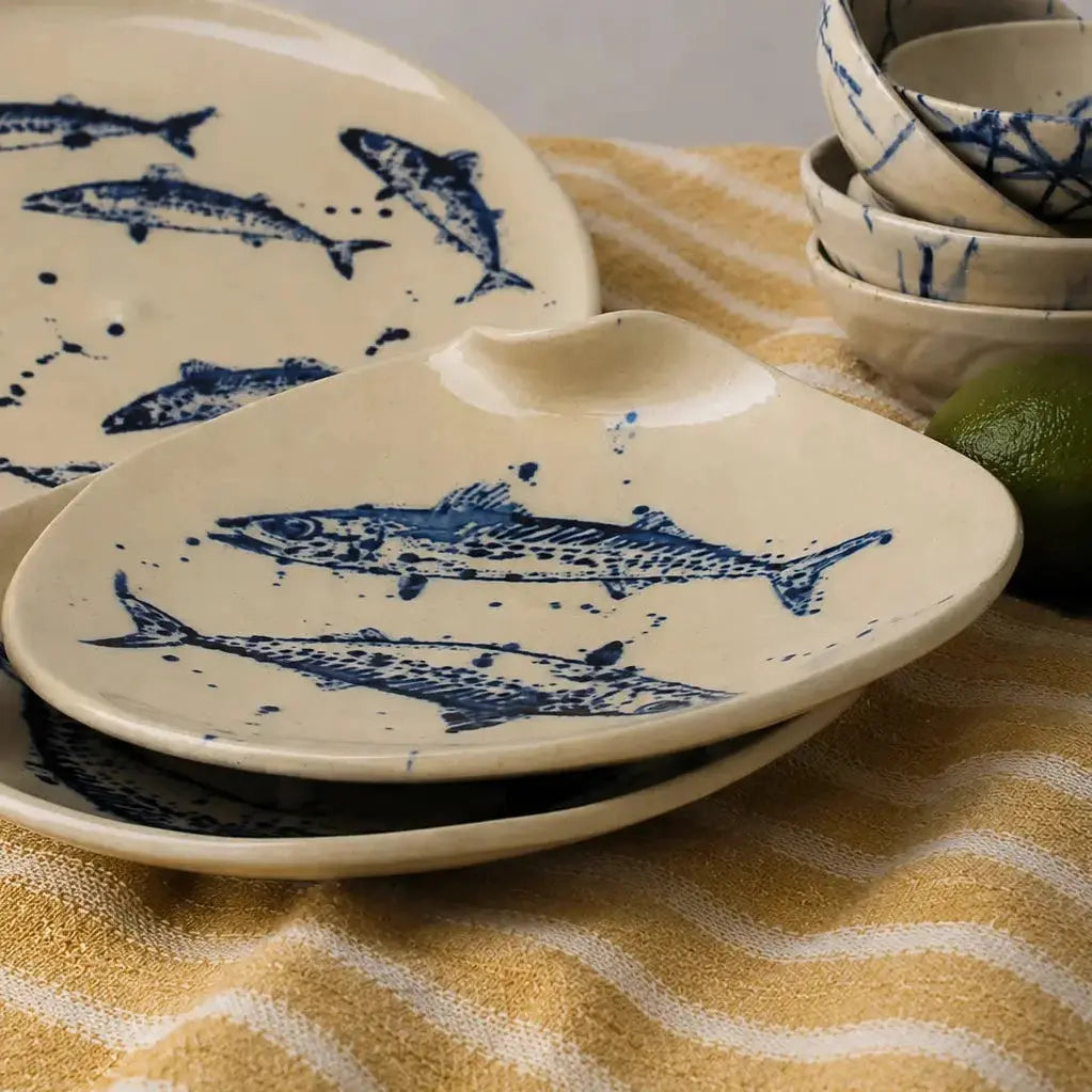 A stack of ceramic fish-shaped plates and bowls with blue fish illustrations, styled on a yellow and white striped cloth with a lime.