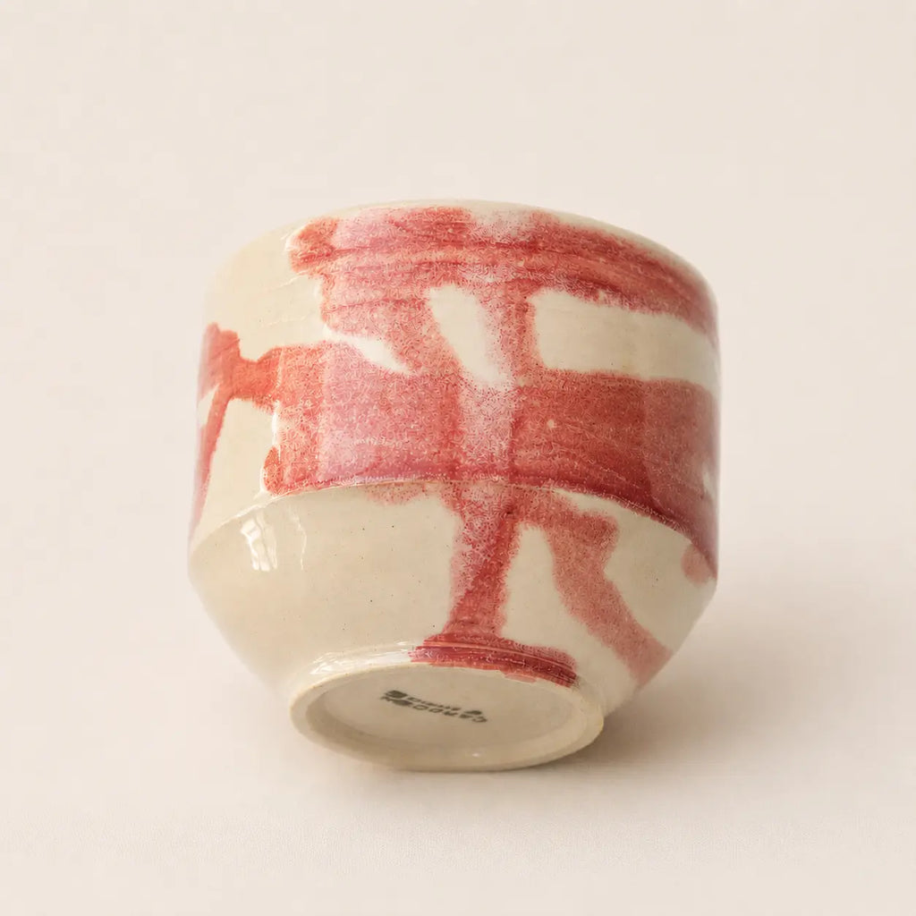 A small ceramic chawan matcha cup with a glossy off-white glaze and a bold red painted brushstroke pattern, shown from a side angle on a light beige background.