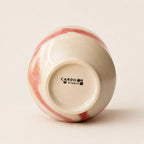 An off-white ceramic chawan matcha cup shown from its base, featuring a glossy finish with pinkish-red marbled swirls and the black printed brand name 'CARDON STUDIO' in the center.