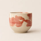 A small ceramic chawan matcha cup with a rounded shape, featuring a glossy off-white glaze and abstract reddish-pink brushstrokes around the exterior, set against a plain light beige background.