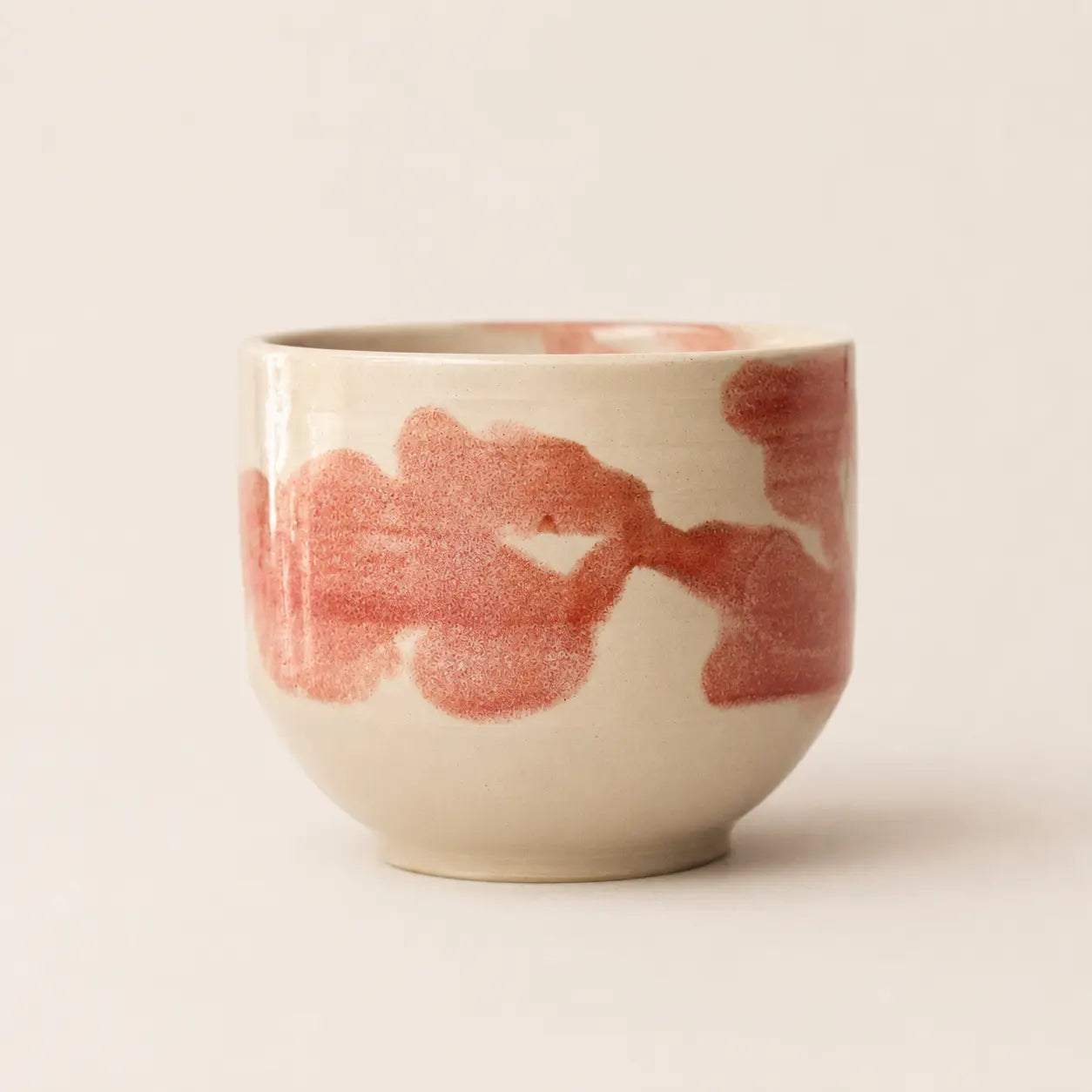 A small ceramic chawan matcha cup with a rounded shape, featuring a glossy off-white glaze and abstract reddish-pink brushstrokes around the exterior, set against a plain light beige background.
