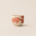 A small ceramic espresso cup with a warm cream base and abstract pink brushstroke patterns, set against a solid, warm off-white background.