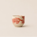 A small ceramic espresso cup with a warm cream base and abstract pink brushstroke patterns, set against a solid, warm off-white background.