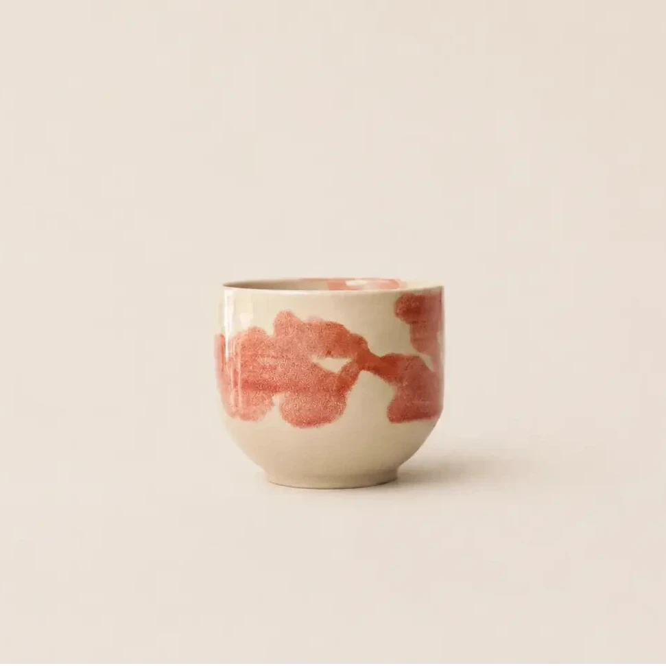 A small ceramic espresso cup with a warm cream base and abstract pink brushstroke patterns, set against a solid, warm off-white background.
