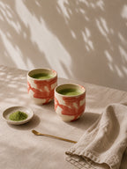 A small ceramic espresso cup with a warm cream base and abstract pink brushstroke patterns, set against a solid, warm off-white background.