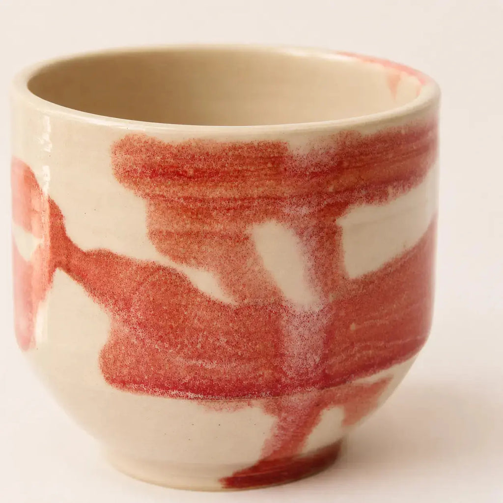 A ceramic chawan matcha cup with a glossy off-white glaze and abstract red painted brushstrokes, photographed against a plain light background.