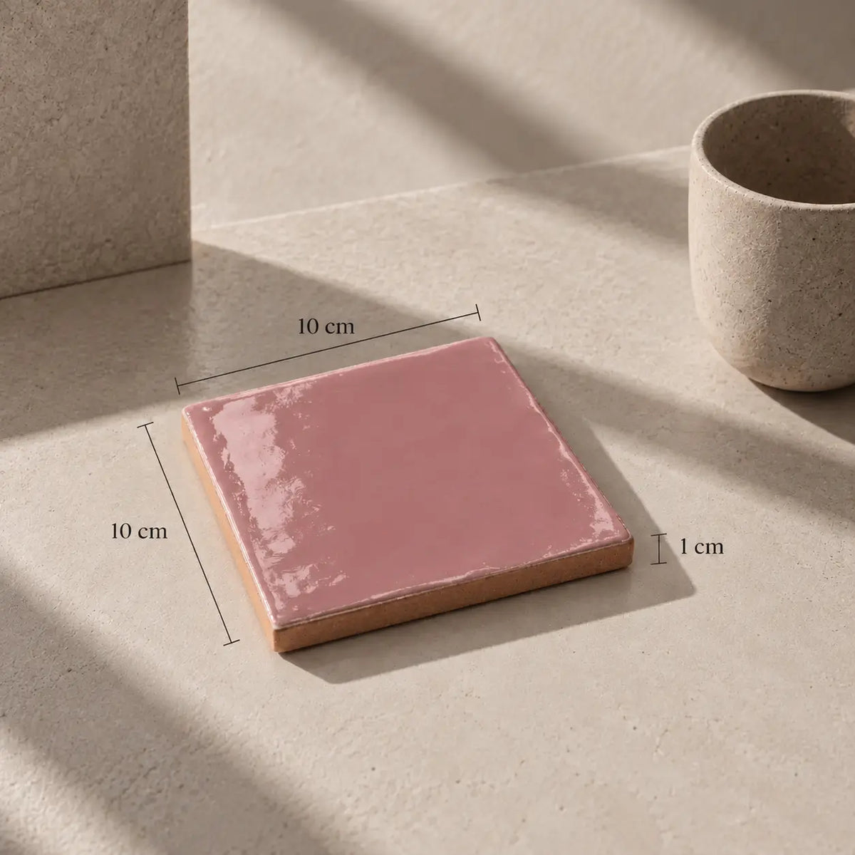 A square pink ceramic tile with a glossy finish and a natural clay edge, shown on a beige stone surface with dimension lines indicating 10 cm by 10 cm and a 1 cm thickness.