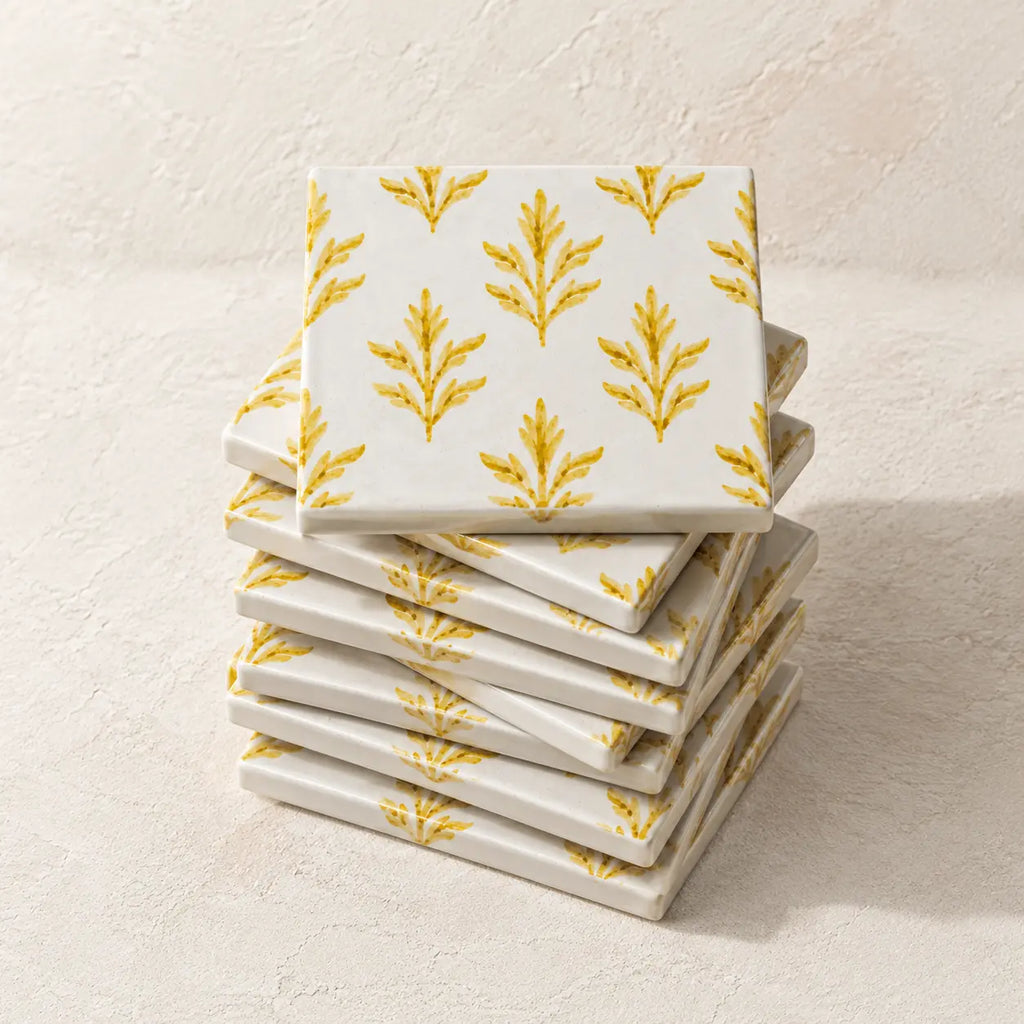 A stack of square white ceramic coasters with a repeating yellow botanical leaf pattern, photographed on a light textured beige surface.