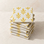 A stack of square white ceramic coasters with a repeating yellow botanical leaf pattern, photographed on a light textured beige surface.