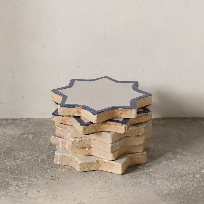 A stack of star-shaped ceramic coasters with a white center and a dark blue border, set on a gray concrete floor against a light gray wall.