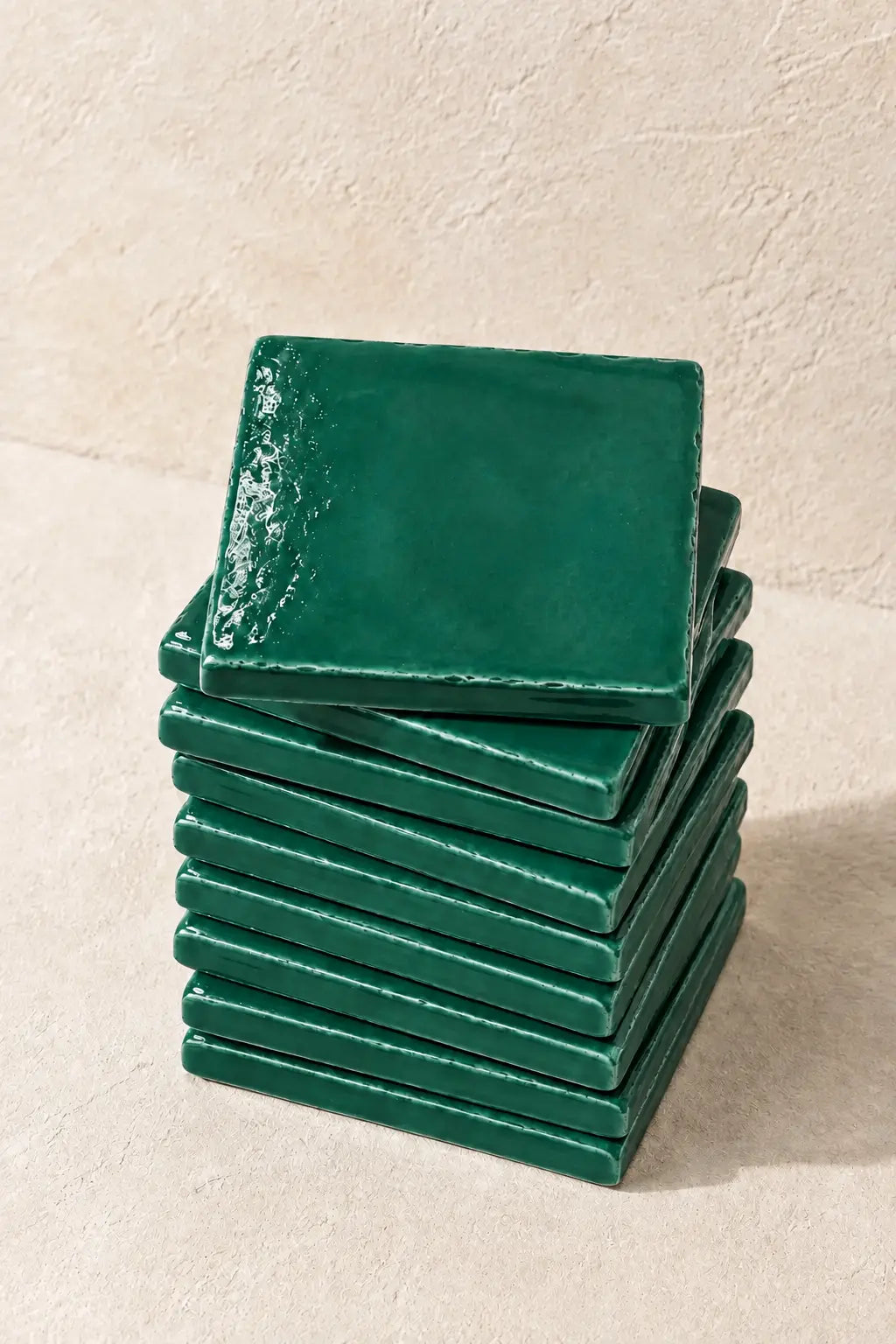 A stack of glossy emerald dark green square coasters on a textured beige surface against a matching beige wall.