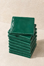A stack of glossy emerald dark green square coasters on a textured beige surface against a matching beige wall.