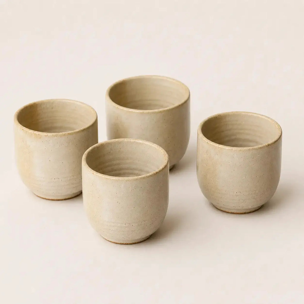 Four small handmade ceramic espresso cup in a light beige stoneware finish, arranged on a plain off-white background.