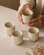 Three small beige ceramic cups on a light fabric table surface, with a person's hands holding one cup filled with tea. A neutral indoor setting and dried flowers are softly visible in the background.